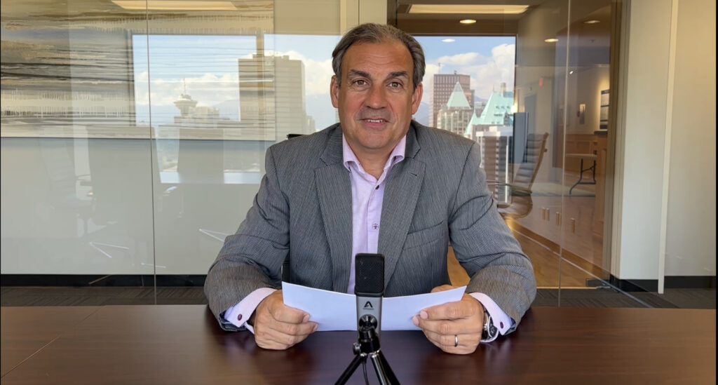 Constantine Lycos sits in a boardroom with notes, a mug, and a microphone on the table in front of him and a reflection of downtown Vancouver in the background