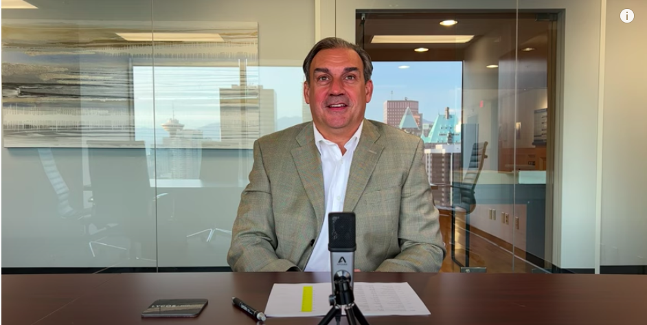 Constantine Lycos sits in a boardroom with notes and a microphone on the table in front of him and a reflection of downtown Vancouver in the background