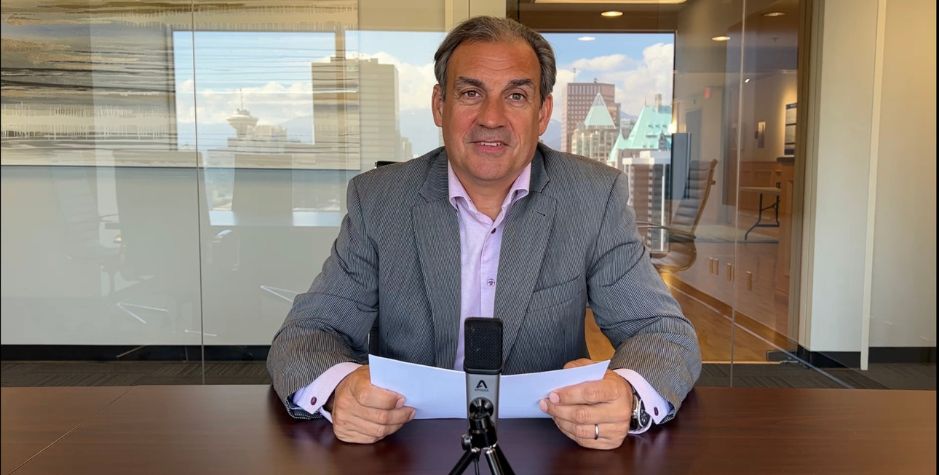 Constantine Lycos sits in a boardroom with notes and a microphone on the table in front of him and a reflection of downtown Vancouver in the background