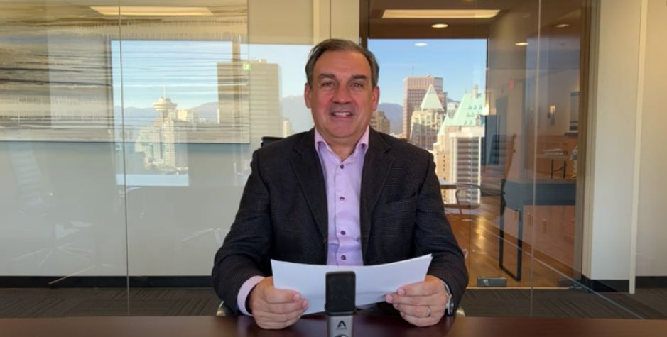 Constantine Lycos sits in a boardroom with notes and a microphone on the table in front of him and a reflection of downtown Vancouver in the background